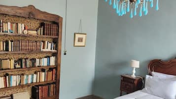 A bedroom with a large wooden bookshelf, a chandelier, a bedside table with a lamp, and a bed with white linens.