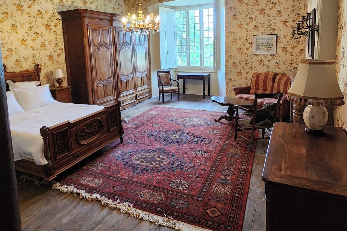 Une chambre à coucher avec un lit à baldaquin, un lustre, une grande fenêtre, une commode en bois et un tapis à motifs.