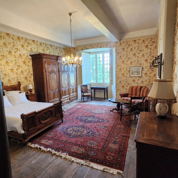 A bedroom with a four-poster bed, a chandelier, a large window, a wooden dresser, and a patterned rug. - Château de Trévien (Trévien)