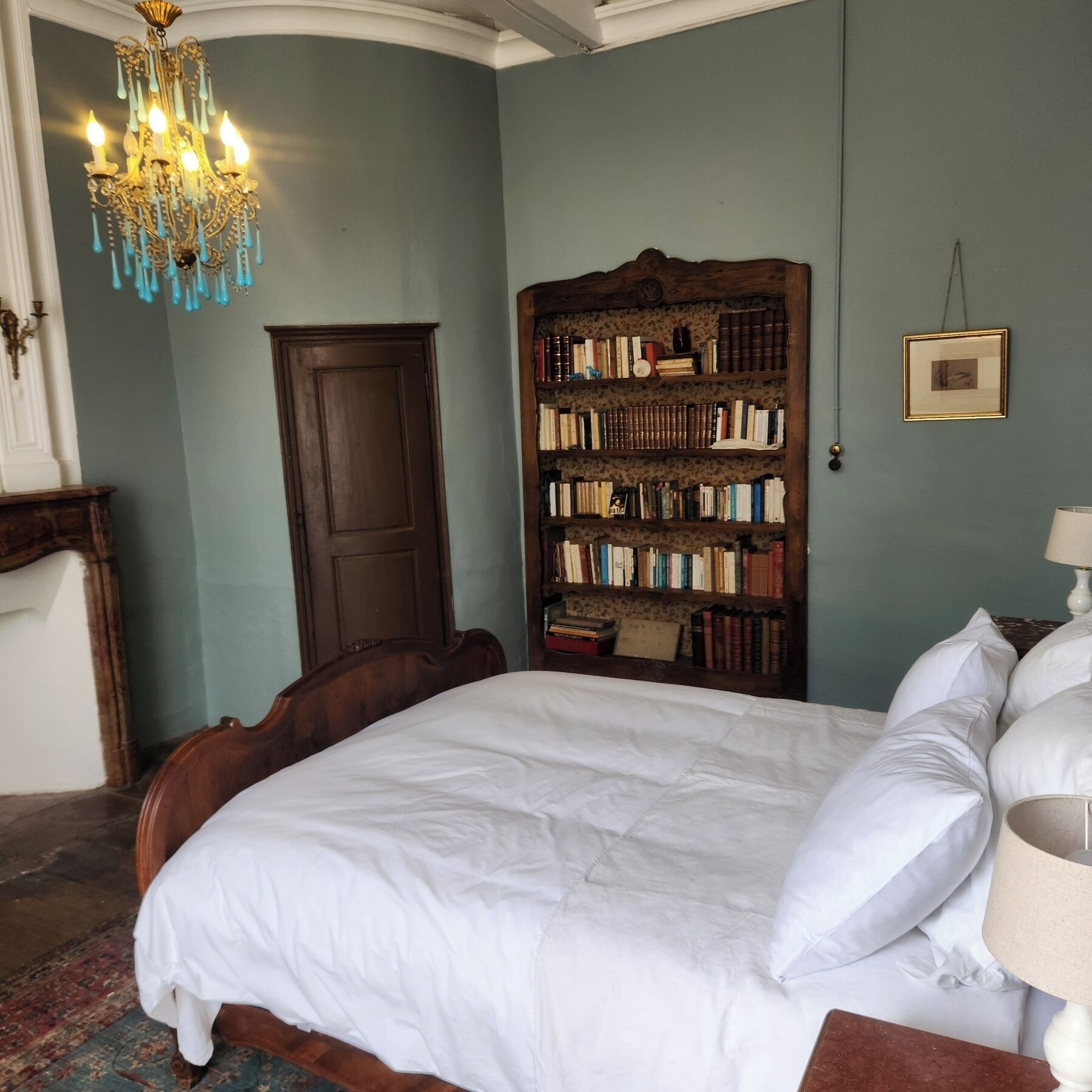 A bedroom with a large bed, a wooden bookshelf, a chandelier, and a door.