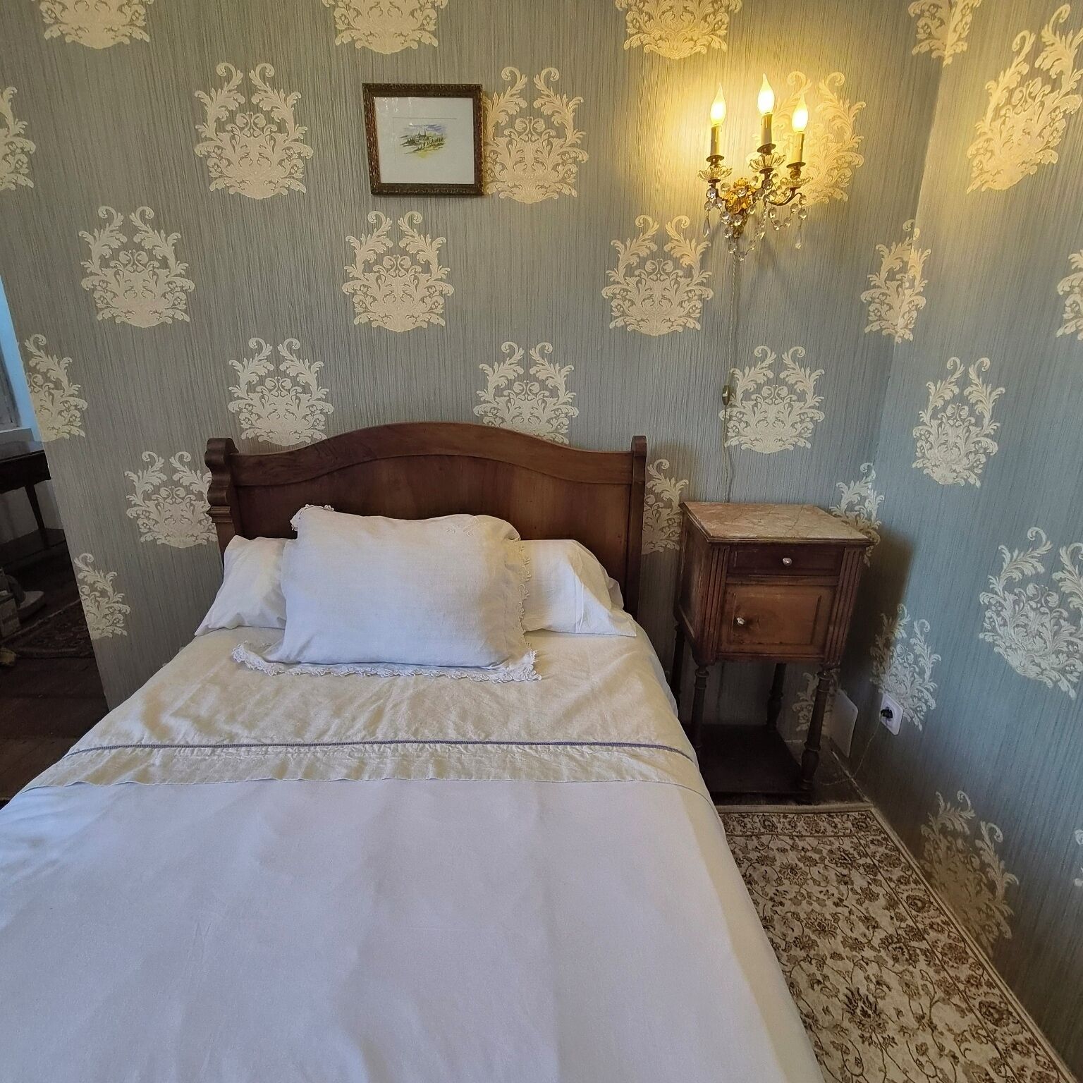 A neatly made bed with white linens, a wooden headboard, a nightstand with a drawer, and a wall-mounted light fixture.