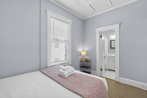 A bedroom with a bed, a bedside lamp, a window with blinds, and a door leading to a bathroom. - Upper Carriage House at Wayne Estate (Savannah)