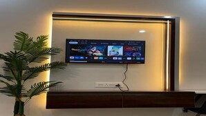 A flat-screen TV mounted on a wall with a wooden cabinet below. - Tranquil studio with AC in enjoyable Greater Noida location (Greater Noida)