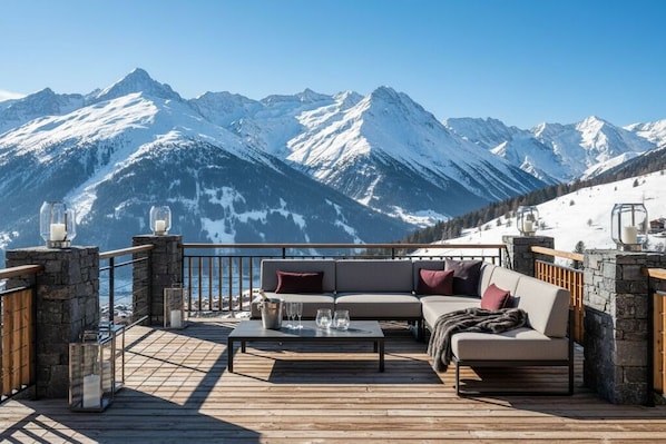 A deck with a sofa and table, overlooking snow-covered mountains.