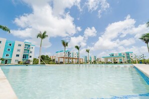 A swimming pool with clear water, surrounded by multi-story buildings with balconies, palm trees, and a clear sky. - Elegant Pool-View Apartment Near Punta Cana Beach (Punta Cana)