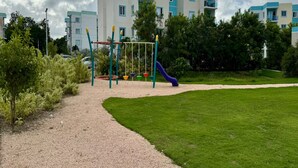 Un'area giochi con altalene e uno scivolo, circondata da un vialetto sterrato e prato verde, con un edificio a più piani sullo sfondo.