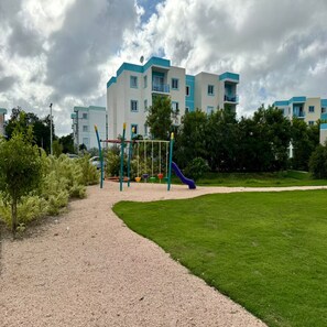 Ein Spielplatz mit Schaukeln und Rutsche, umgeben von einem Kiesweg und grünem Gras, mit einem mehrstöckigen Gebäude im Hintergrund.
