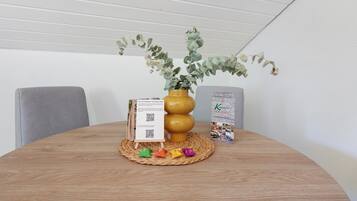A round wooden table with a woven placemat, a yellow vase with green foliage, a QR code stand, and a brochure.
