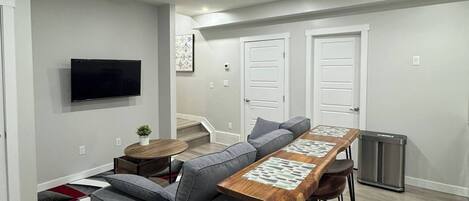 A modern living room with a sectional sofa, a wooden dining table with bar stools, a flat-screen TV mounted on the wall, and a staircase leading to an upper level.