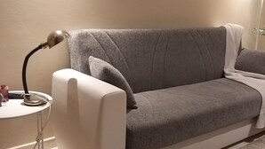 Ett vardagsrum med en grå soffa, ett litet sidobord med en lampa, en klocka på väggen och ett golv täckt av en matta.