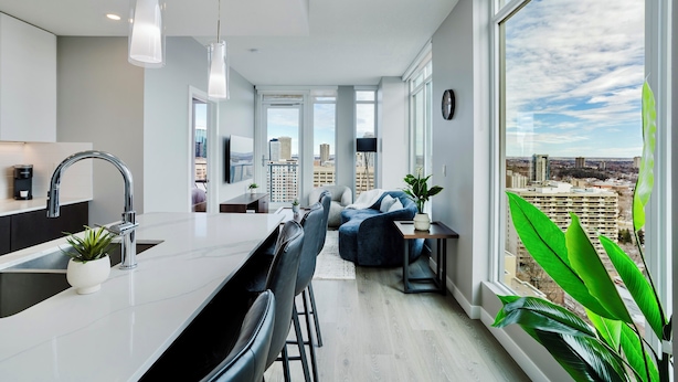 A modern kitchen with a large island, a dining area with chairs, and a view of the city through large windows. - Epic River Valley View | 20th floor | Near Rogers (Edmonton)
