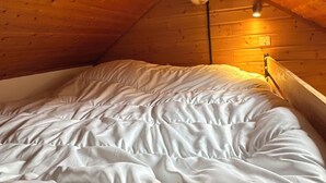 Un llit individual en una habitació de cabana de fusta amb sostre inclinat i una lluminària penjada a la paret.
