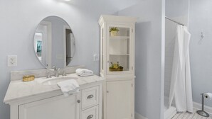 A bathroom with a white vanity, a large mirror, a cabinet with glass doors, a shower with a white curtain, and a wall-mounted light fixture. - Lower Carriage House at Wayne Mansion Estate (Savannah)