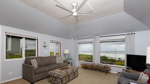 A living room with a sofa, ottoman, TV, and large windows overlooking the ocean.