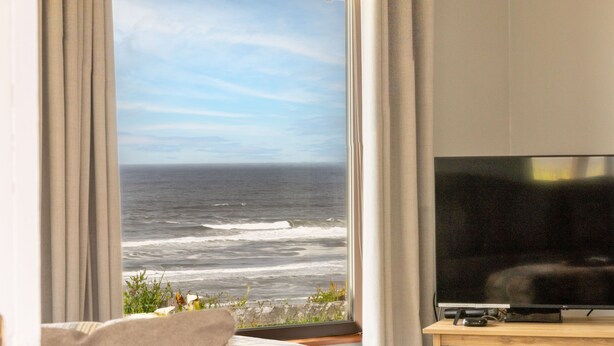 A room with a large window overlooking the ocean, a flat-screen TV on a wooden stand, and a curtain partially drawn.