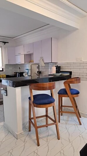 A kitchen with a marble floor, white cabinets, a black countertop, and two wooden bar stools with blue cushions.