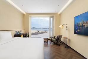A hotel room with a bed, a desk, a chair, a lamp, and a large window with a view of the beach. - Bay Four Seasons Castle Hotel (Weihai)