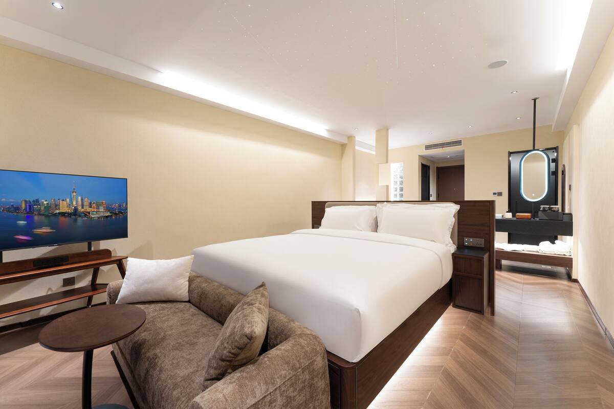 A hotel room with a large bed, a TV, a sofa, a small table, a mirror, and a wooden floor.