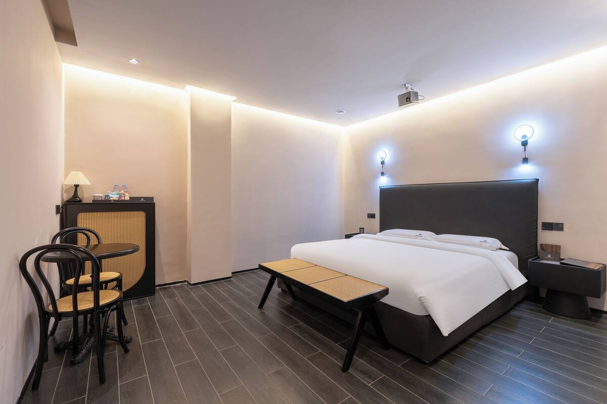 A modern hotel room with a bed, bedside tables, a desk with chairs, and a bench.