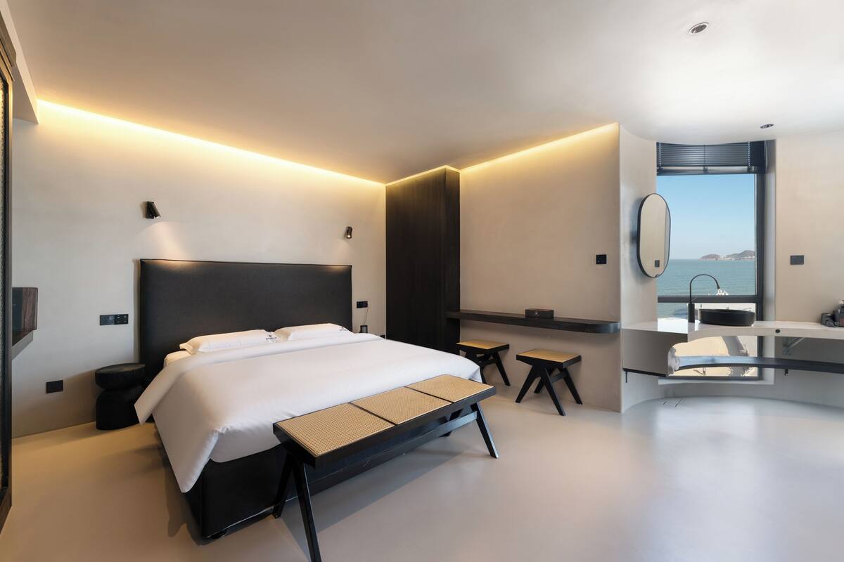 A modern hotel room with a bed, bedside table, bench, and a view of the sea through a window.