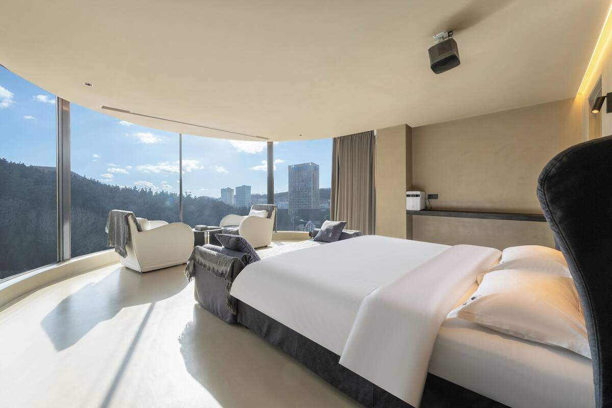 A modern hotel room with a large bed, a seating area, and a view of the cityscape.