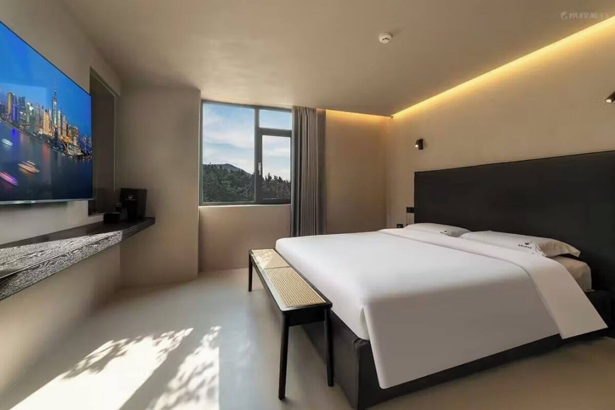 A modern hotel room with a large bed, a television mounted on the wall, a window with a view, and a bench.