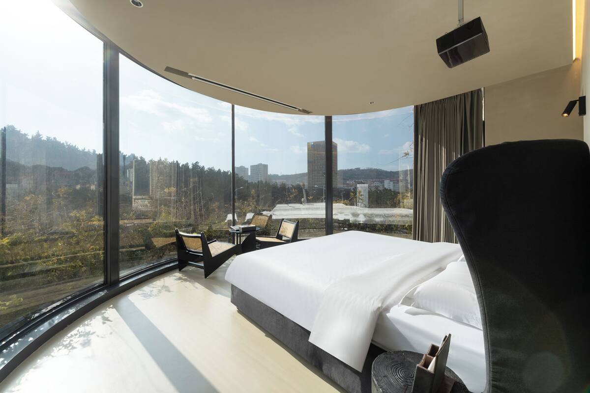 A modern hotel room with a large bed, a curved ceiling, and a panoramic view of a cityscape and forest.