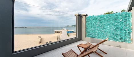 A balcony with a view of the beach, a wooden chair, and a small table.
