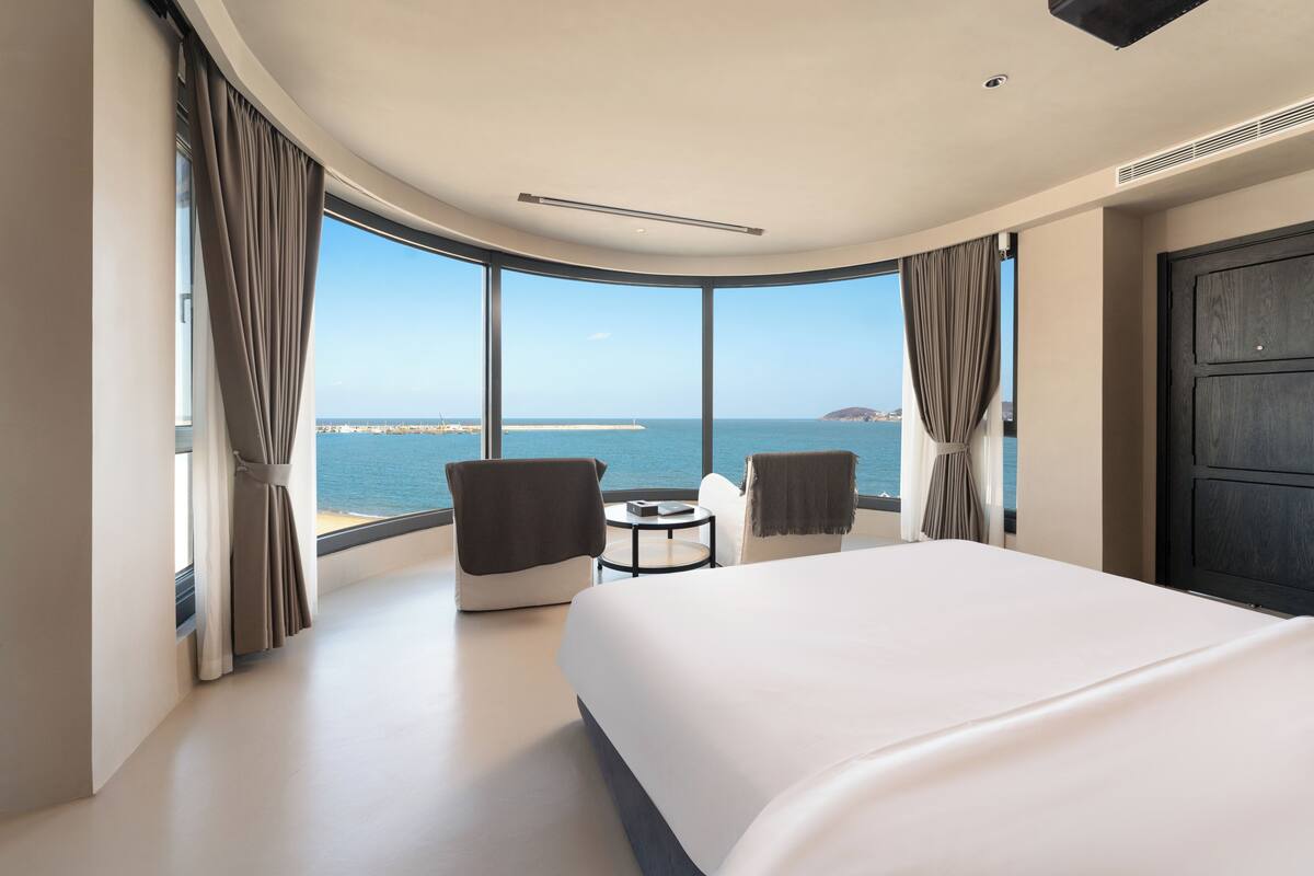 A modern hotel room with a large bed, two armchairs, a small table, and a view of the sea.