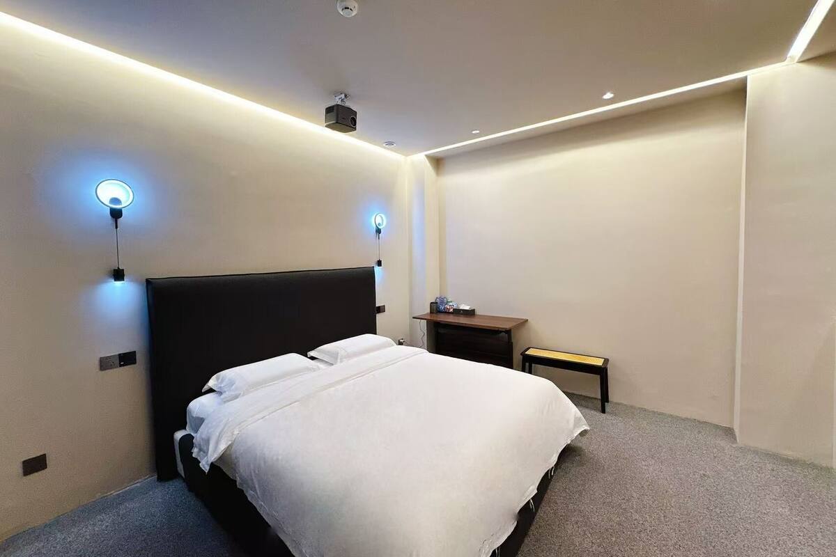 A modern hotel room with a bed, bedside lamps, a projector, and a small table.