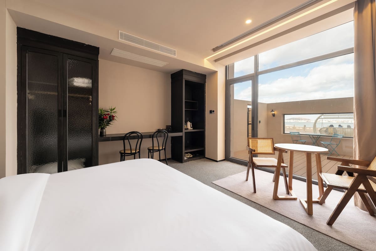 A hotel room with a large bed, a desk with chairs, a bookshelf, and a view of the city through large windows.