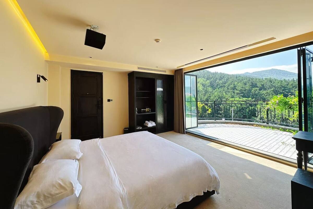 A bedroom with a large bed, a wardrobe, a seating area, and a balcony with a view of a forested landscape.