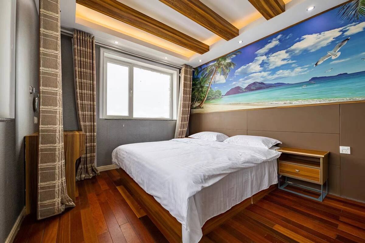 A bedroom with a bed, a nightstand, a window, and a beach mural on the wall.