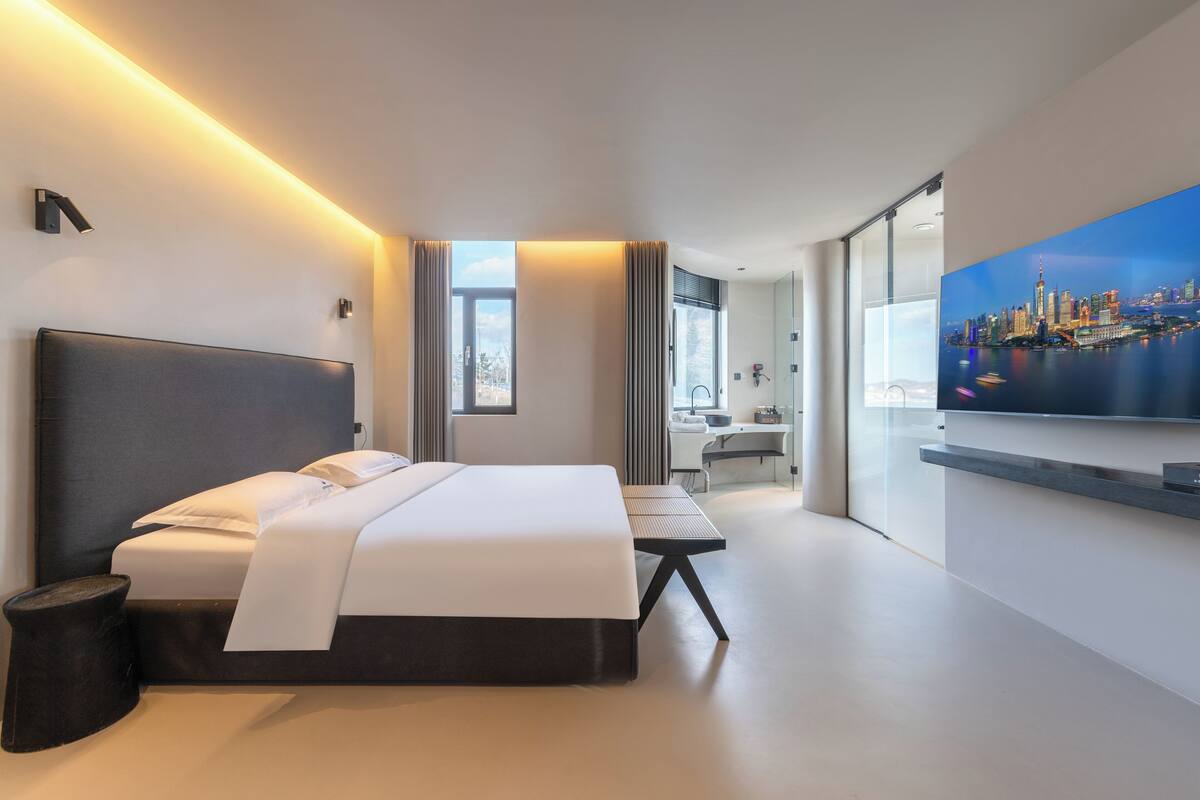 A modern hotel room with a large bed, a desk, a chair, a TV, and a cityscape view.