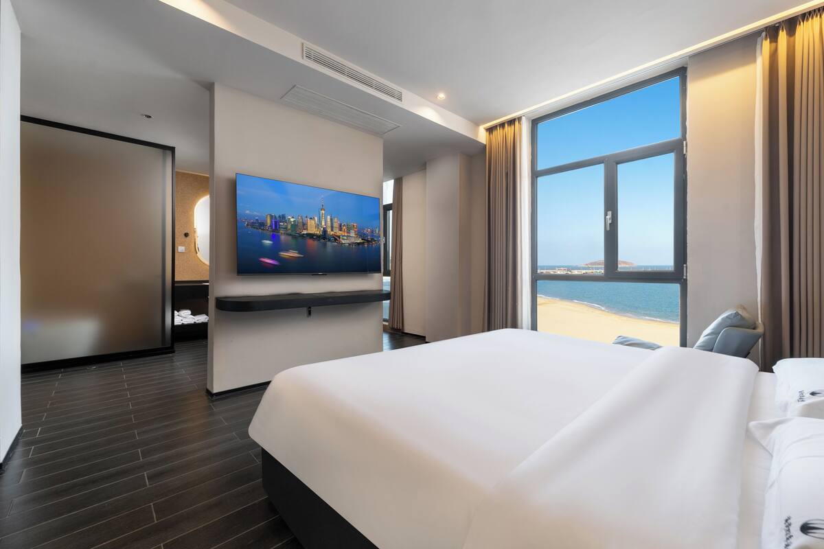 A hotel room with a large bed, a flat-screen TV, a cityscape view, and a large window.