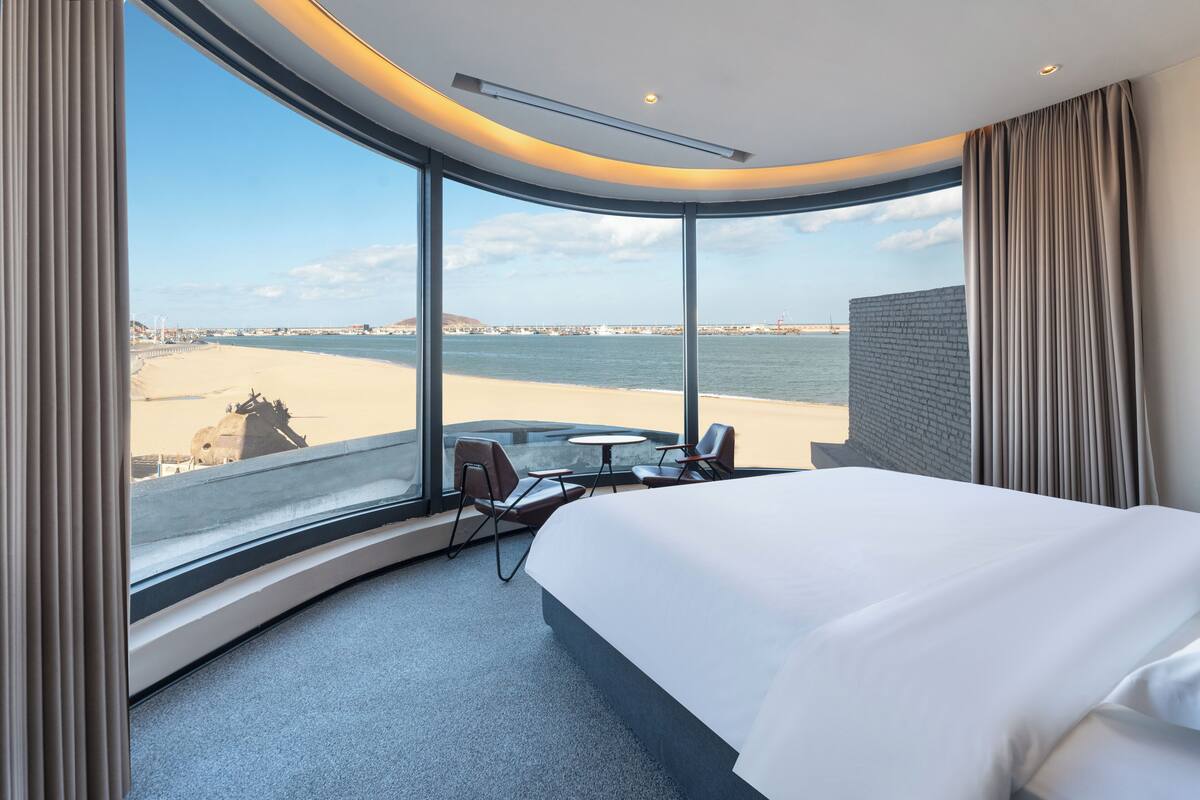 A hotel room with a curved window, a bed, a small table, and two chairs.