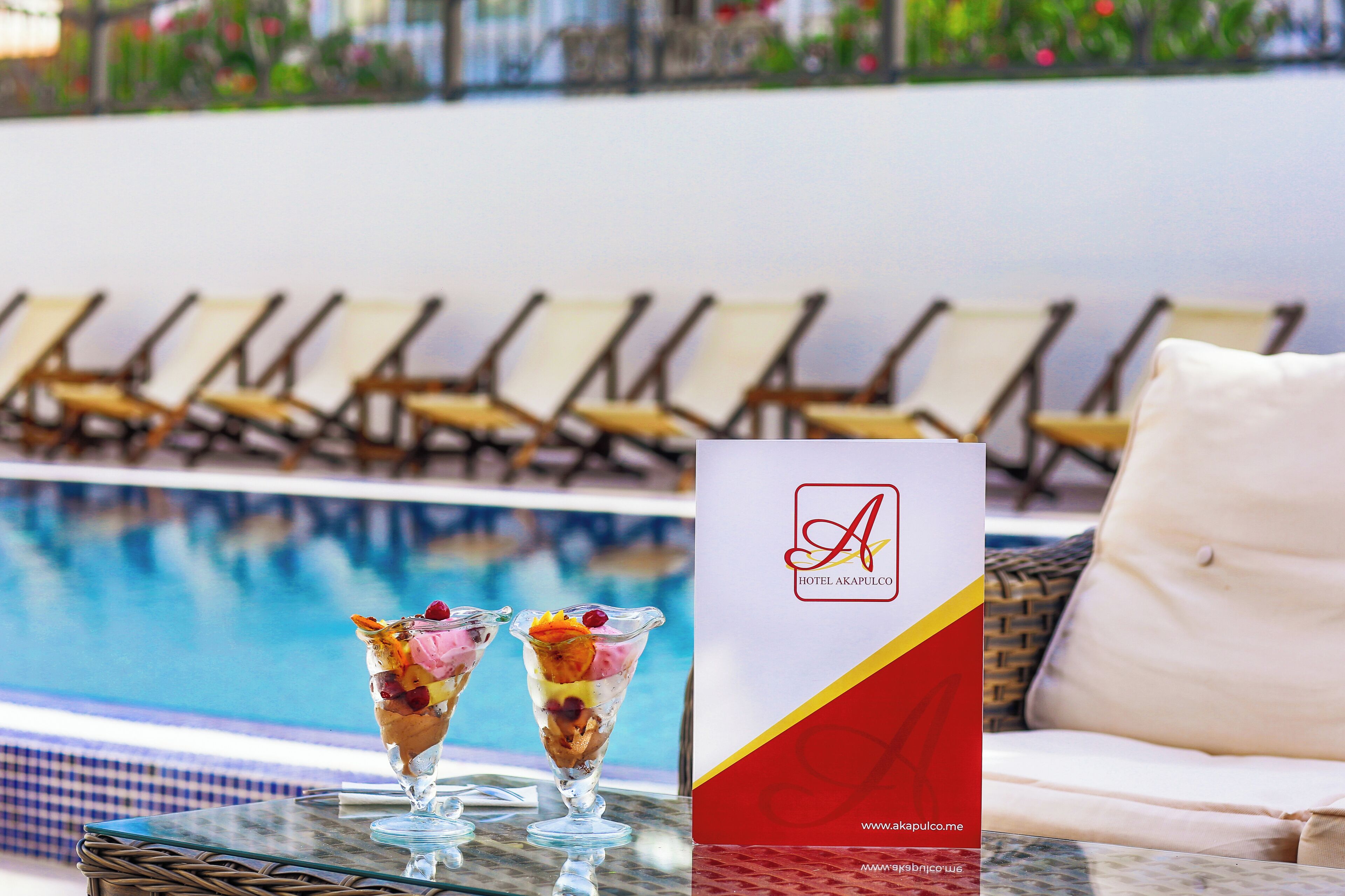 A poolside setting with lounge chairs, a glass table with two dessert drinks, and a hotel brochure.
