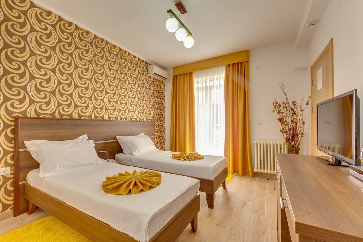 A hotel room with two beds, a TV, a wooden desk, and a window with curtains.