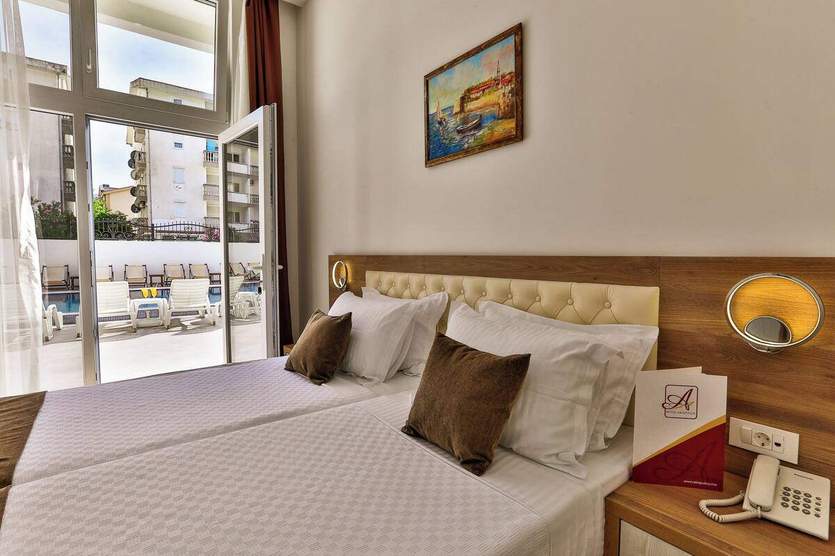 A hotel room with a bed, a bedside table with a phone, a painting on the wall, and a view of the outside.