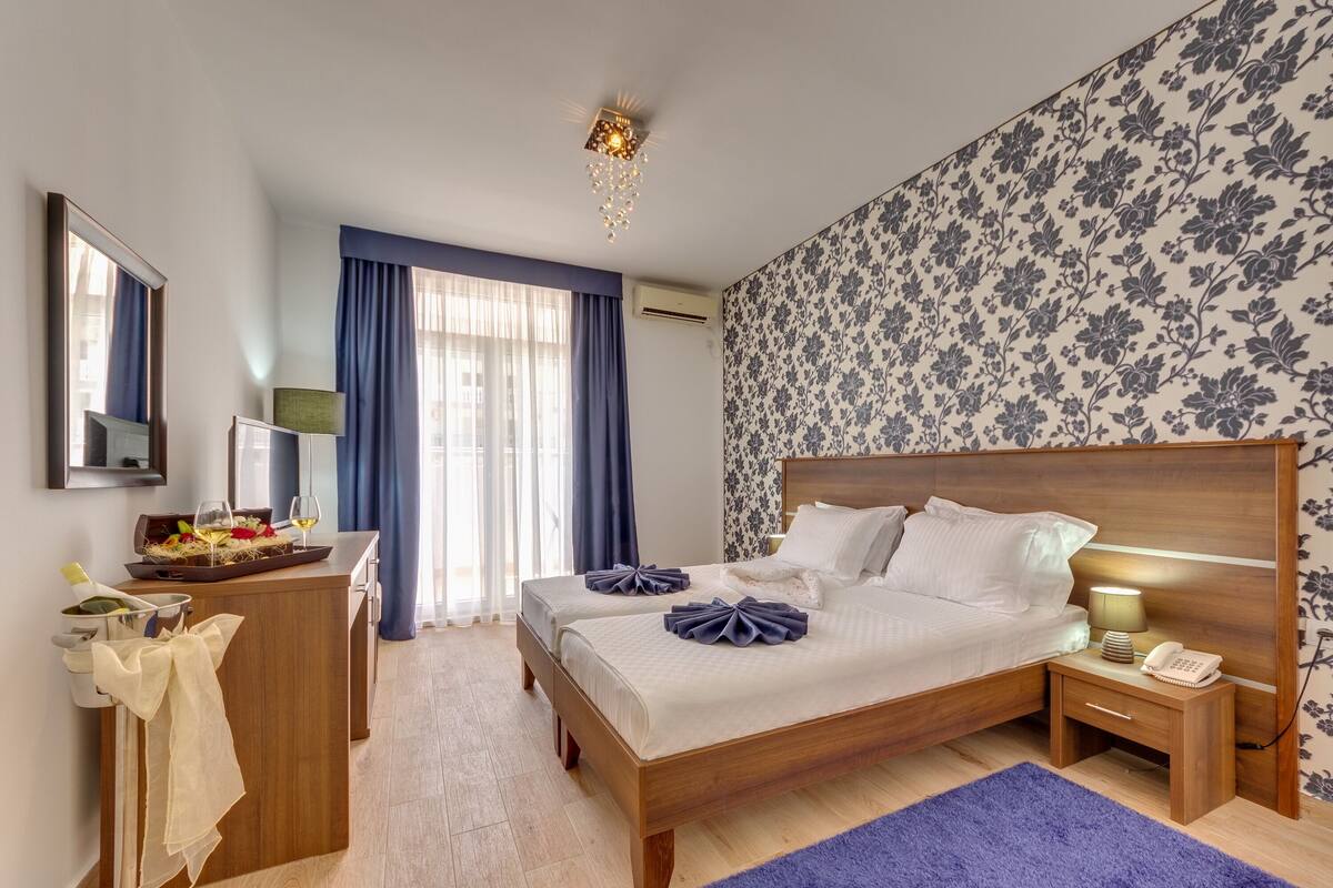 A hotel room with a wooden bed, bedside tables, a desk with a lamp, and a large window with curtains.