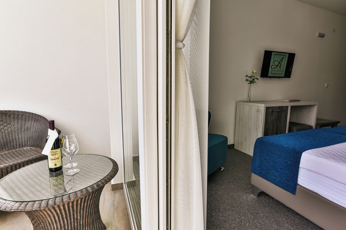 Quarto de hotel com mesa de vidro, cadeira de vime, cama com cobre azul, pequeno armário e quadro na parede.
