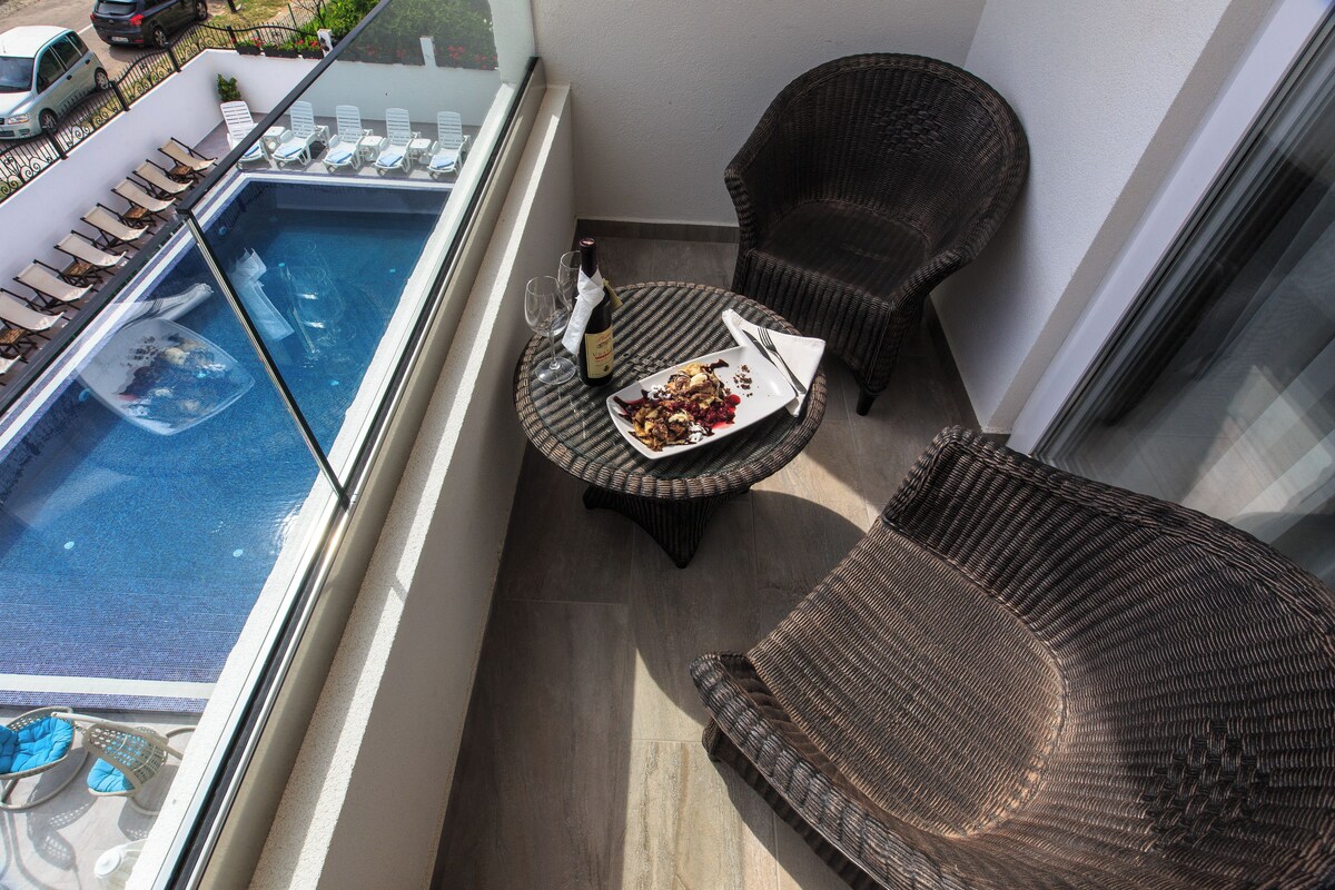 A balcony with a pool, wicker chairs, a dining table with a bottle and glasses, and a plate of food.