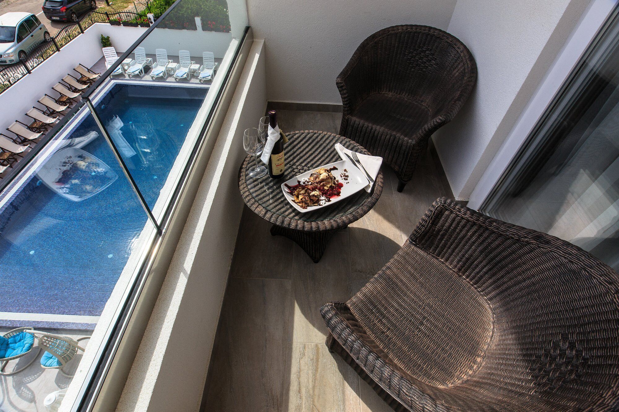 A balcony with a pool, lounge chairs, a wicker chair, and a table with a plate of food and a wine glass.