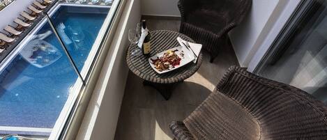 A balcony with a pool, lounge chairs, a wicker chair, and a table with a plate of food and a wine glass.