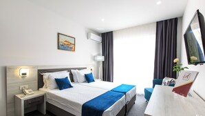A hotel room with a bed, bedside tables, a desk, a chair, an air conditioning unit, a painting, and a window with curtains.