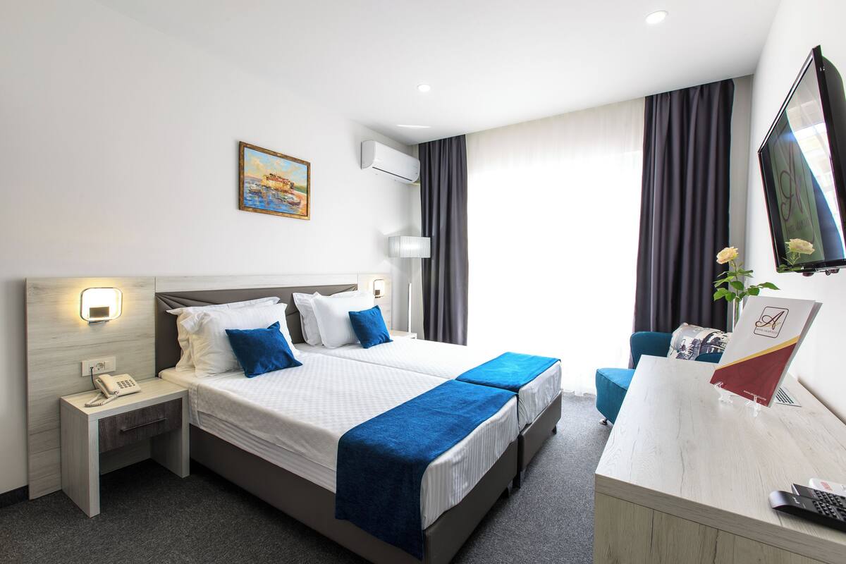 A hotel room with a bed, bedside tables, a desk, a chair, an air conditioning unit, a painting, and a window with curtains.