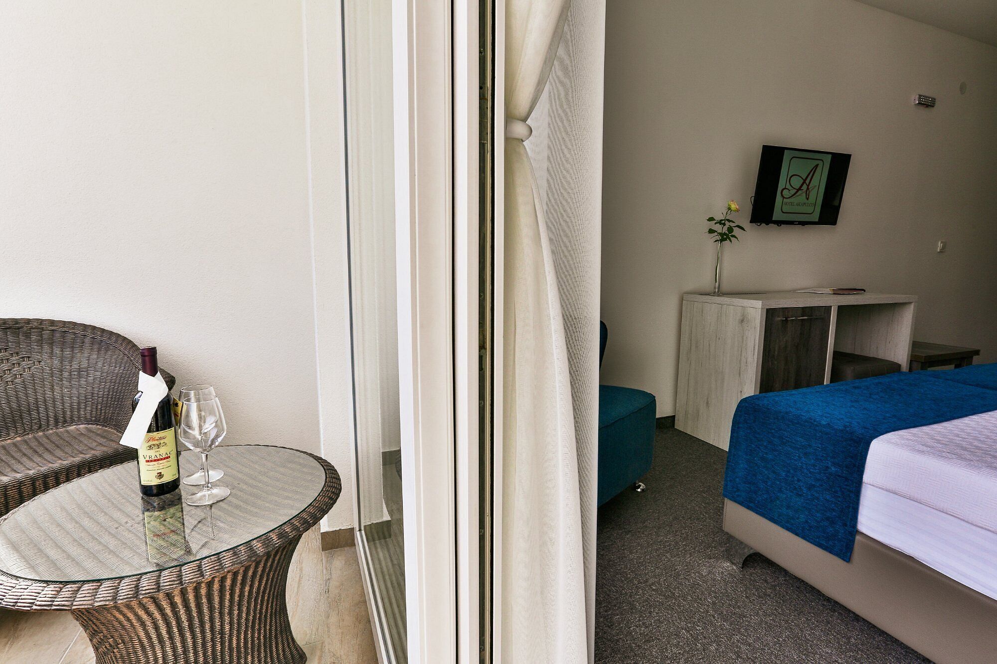 A hotel room with a wicker chair, a small glass table with a bottle and glass, a bed with a blue cover, a desk with a vase, and a framed picture on the wall.