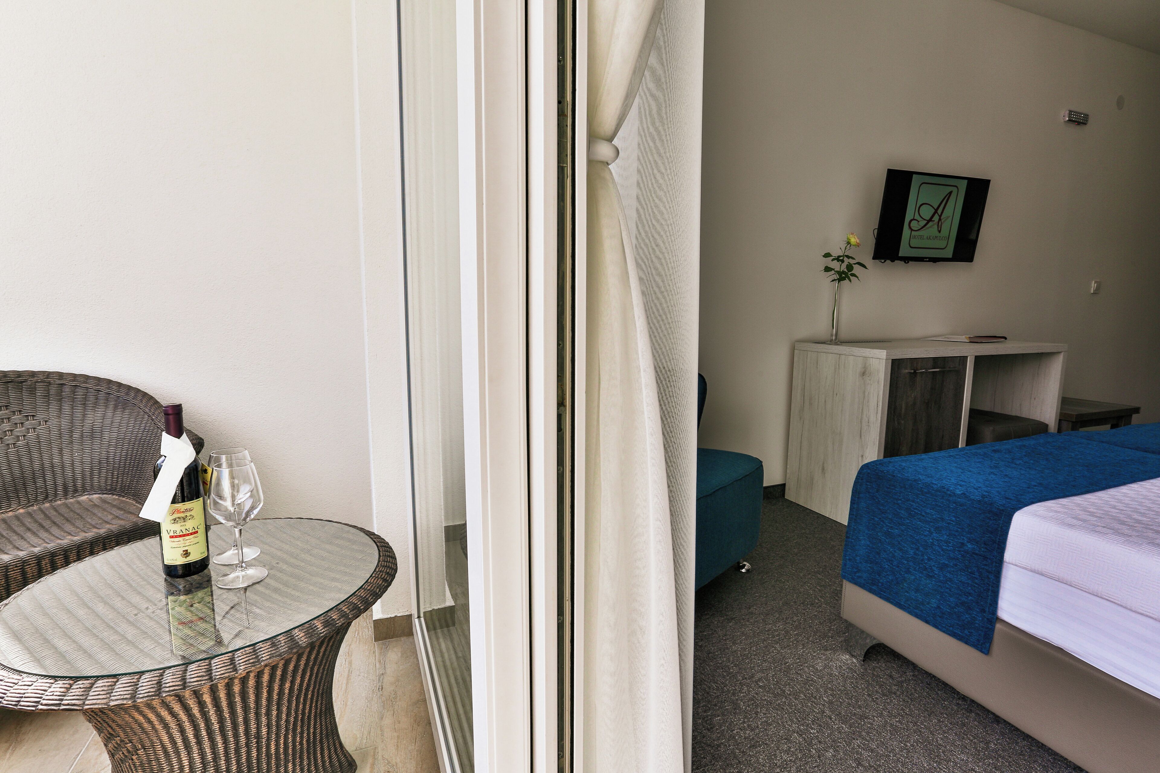 Una habitación de hotel con una mesa de vidrio, una silla de mimbre, una cama con cubrecama azul, un pequeño armario y un cuadro en la pared.