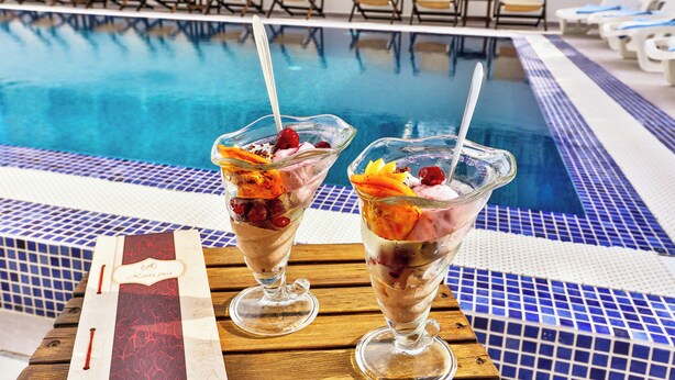 Two dessert glasses on a wooden table by a poolside with lounge chairs in the background.