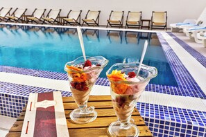 Two dessert glasses on a wooden table by a poolside with lounge chairs in the background.